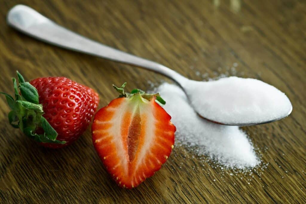 A pair of strawberries next to a spoonful of sugar.
