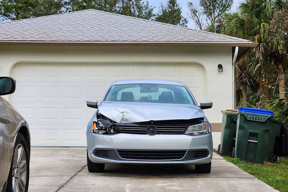 A grey car in the driveway with collision damage to the right front headlight and hood.